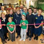Group of people behind a ribbon. There is a new 'Optometry' wall design in the background. One person wears a pink 'Oxford Hospitals Charity' t-shirt.
