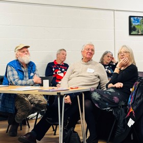 Five older people wearing colourful clothes, seated on chairs and at a table, listening intently.