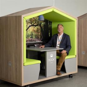 A smiling man in a jacket and rainbow lanyard sits on a padded bench at a small table, an integral part of a mini hut on wheels.