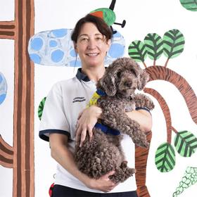 A young woman in a white T-shirt holds in her arms a relaxed curly-haired floppy-eared dog. On the wall behind are naïve stylised images of branches and leaves.