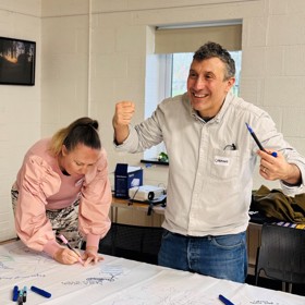 A young women in a bright shirt draws carefully on the corner of a sheet on a table, while an expressive man with a 'Carmine' name sticker makes a cheerful gesture to someone watching.