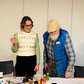 An older man and younger woman stand a table covered with a large sheet of paper and written messages. Paints and pens are scattered on the table.