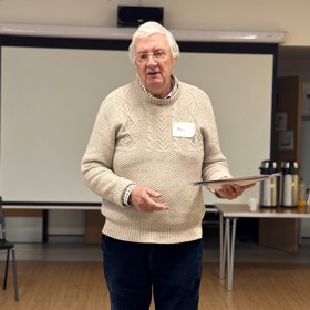 An older man standing with some papers in his hand, speaking; table of coffee urns behind.