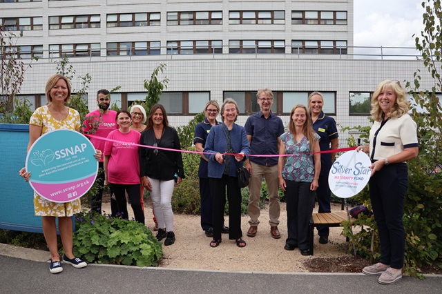 Eleven people in the new garden. One person is about to cut the ribbon. Two people hold banners for 'Silver Star Maternity Fund' and 'SSNAP'