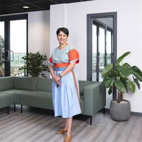 An elegant woman in a smart, brightly coloured summer dress stands in a light modern space with a sofa and large indoor plants; tall glass doors on both sides of her lead out to a high terrace with fields in the distance.