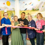 Group of seven smiling people. A couple are cutting a pink ribbon and two people are wearing pink 'Oxford Hospitals Charity' t-shirts.
