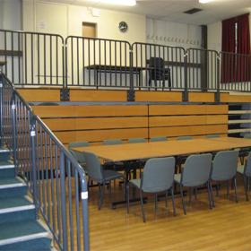 Split level hall with steps up in foreground, polished floor: long table with six seats each side on lower level, railings along steps and upper level.
