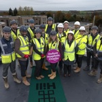 Group of 15 people on a roof and wearing PPE (helmets and high visisiubility jackets). One person holds a banner with the text 'Oxford Hospitals Charity'). Completed buildings are in the background.