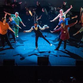 Tap-dancing man on stage with three dancers in NHS scrubs on either side