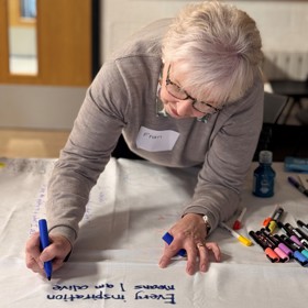 An older woman with a 'Fran' name sticker writes on a white sheet: ' Every inspiration means I am alive'.