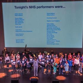 Crowd of NHS staff, some in unform, stand on a stage and receive applause; screen behind them displays their names