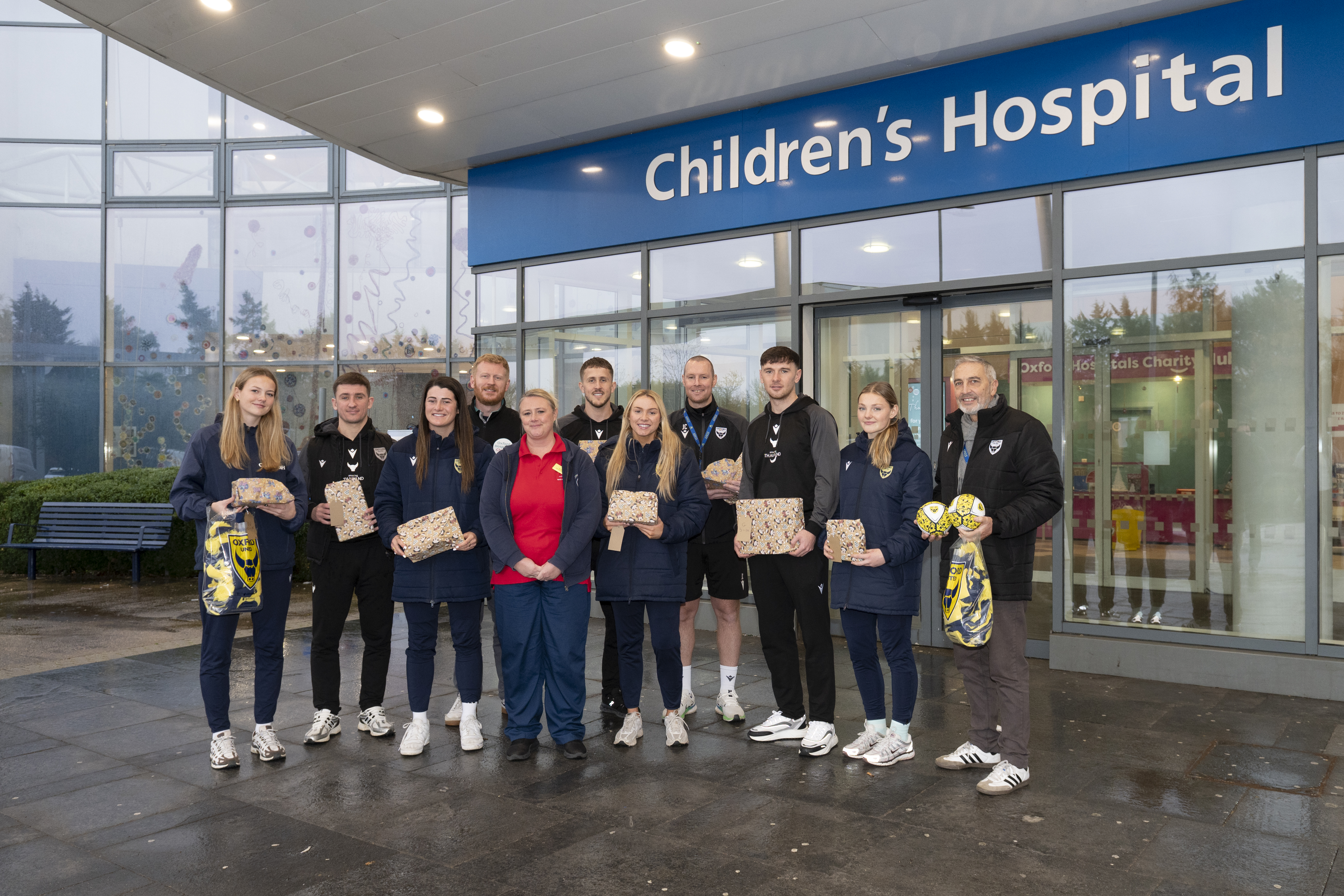 Players and staff from Oxford United, wearing club tracksuits, hold Christmas presents. Other members of OUH and Oxford Hospitals Charity staff are with them in front of the "Children's Hospital" entrance.
