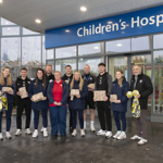 Players and staff from Oxford United, wearing club tracksuits, hold Christmas presents. Other members of OUH and Oxford Hospitals Charity staff are with them in front of the "Children's Hospital" entrance.