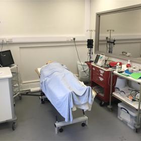 Mannekin on hospital trolley, covered with loose blanket, in clinical setting, shelves of supplies under large mirror to one side.