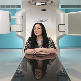 A smiling long-haired woman wearing a patterned blouse and glasses rests her folded arms on the shiny table of a large clinical machine behind her.