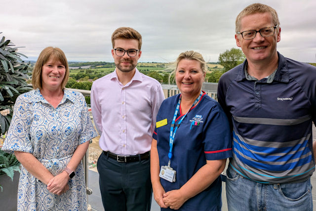 Two women and two men, one woman in nurse uniform, stand smiling with a view of green fields behind