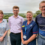 Two women and two men, one woman in nurse uniform, stand smiling with a view of green fields behind
