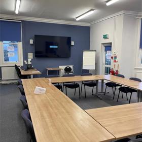 Smooth-top tables arranged in a square, four chairs each side, large digital screen on back wall, flip-pad on stand, two windows, radiator, fire extinguishers on wall.