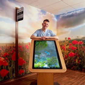 A young man in NHS uniform rests his elbows on a table-sized touch screen tablet. The corner of the room behind is a mural of poppies, sky and clouds, with a fake bus stop on the wall.