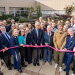 Big group of people, including uniformed members of staff and people wearing 'Oxford Hospitals Charity' t-shirts. Many are smiling. People at the front are holding a pink ribbon and a person is about to cut it.
