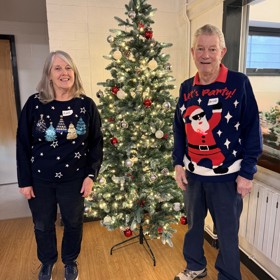 An artificial decorated Christmas tree; standing on its left a woman in a jumper with shiny Christmas tree motifs, and on its right a man in a jumper with Santa wearing shades and 'Let's Party!'.