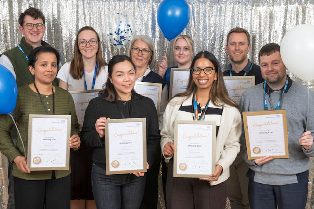 Nine men and women hold up framed certificates with a silver curtain behind; two hold balloons.