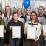 Nine men and women hold up framed certificates with a silver curtain behind; two hold balloons.