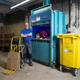 A smiling man in a T-shirt, arms folded, stands in an industrial area alongside a tall metal box: cardboard boxes are seen through a front opening. On one side of him is a trolley with flattened boxes on it and on the other a large plastic wheely bin.