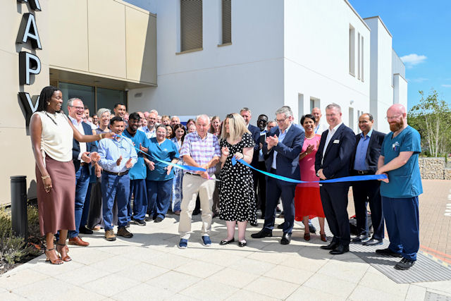 Around 35 smiling people surround two people cutting a ribbon on a smart new paved area on a sunny day