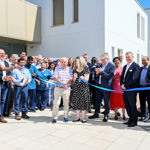Around 35 smiling people surround two people cutting a ribbon on a smart new paved area on a sunny day