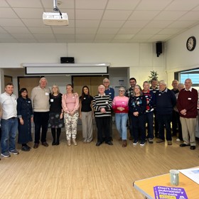 Diverse group of 20 people standing and smiling in a seminar room, Christmas tree behind.