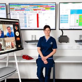 In an office with three large digital screens behind her, a young woman in nurses’ uniform sits alongside a monitor where images of her colleagues can be seen.