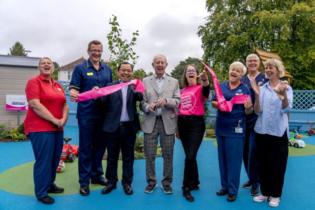 Eight cheerful people, in suits or hospital uniforms, stand in a children's playground and hold up a ribbon