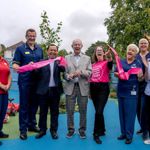 Eight cheerful people, in suits or hospital uniforms, stand in a children's playground and hold up a ribbon