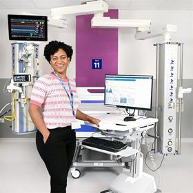 A young woman wearing a lanyard rests her hand on a high-tech digital workstation with a screen - a clean, unoccupied clinical bed bay is behind her.
