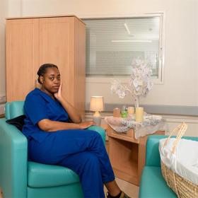 A young woman in blue scrubs sits in a modern armchair in a small calm room. A cabinet next to her bears candles and a model tree with paper hearts attached. An empty Moses basket rests on an armchair on the other side.
