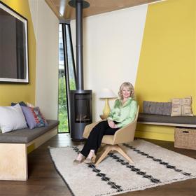 An elegantly dressed woman relaxes in an armchair in a modern, stylish space with rugs and cushioned benches alongside a woodburning stove. Greenery is glimpsed behind through a tall narrow window.
