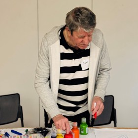 An older man in a striped top has drawn the outline of a balloon in felt tip on the corner of a sheet on a table, and is selecting paint.