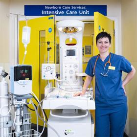 Smiling young woman in blue scrubs with stethoscope around her neck stands in front of bright yellow double doors, leaning on a large, gleaming high-tech machine with controls and monitors.