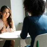 Two women speak to each other across a table in an office setting