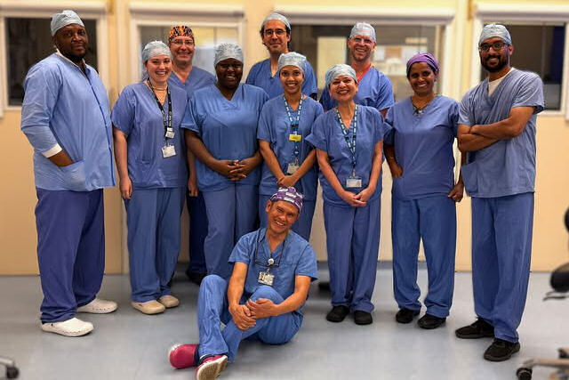 A group of 11 smiling people wearing theatre scrubs in a clinical area