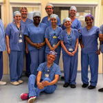 A group of 11 smiling people wearing theatre scrubs in a clinical area