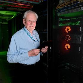 Smiling older man in a smart shirt and lanyard, holding an electronic tablet, stands in a dark room with the glowing controls of computer servers to each side of him.