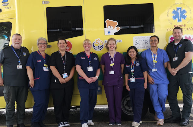Eight people in healthcare uniforms standing in front of an ambulance with a teddy bear picture on it