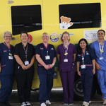 Eight people in healthcare uniforms standing in front of an ambulance with a teddy bear picture on it