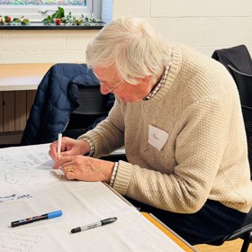 An older man with a name sticker on his jumper sits carefully drawing on the corner of a large sheet of paper.