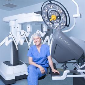 A smiling woman in surgical scrubs sits in a relaxed posture in a clean modern space, with a large items of clinical equipment around her.