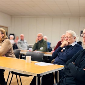 A view of the room with around ten people seated at long tables listening intently.