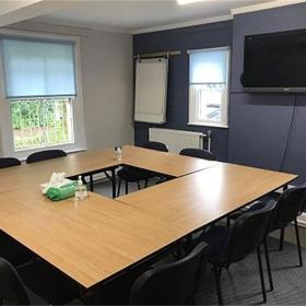 Neat smooth-top tables arranged in a square, chairs tucked under; digital screen, two windows and flip pad on walls.