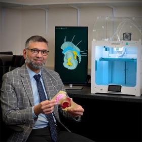 A cheerful man in jacket, tie and glasses holds out a model of a human bone. Behind him there is a medical diagram on a screen and box-shaped 3D printer.